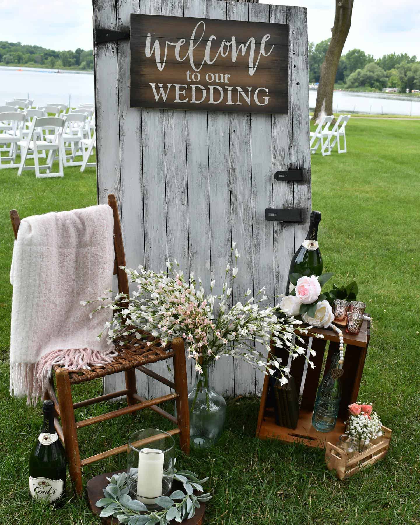 enhancements Outdoor wedding welcome display with a rustic wooden door sign, chair with blanket, flowers, candles, crates, and champagne bottles on grass, with chairs and a lake behind.