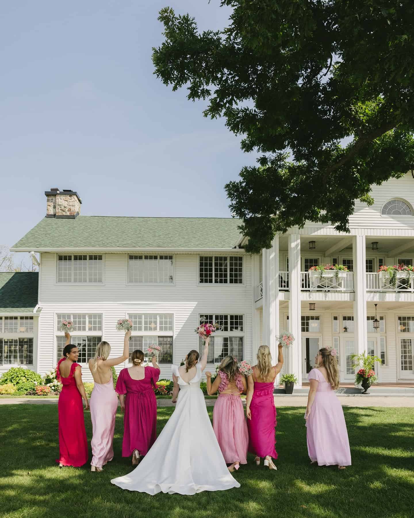upcoming-event A bride in a white gown walks on grass with six bridesmaids in pink dresses, holding bouquets, in front of a large white house.