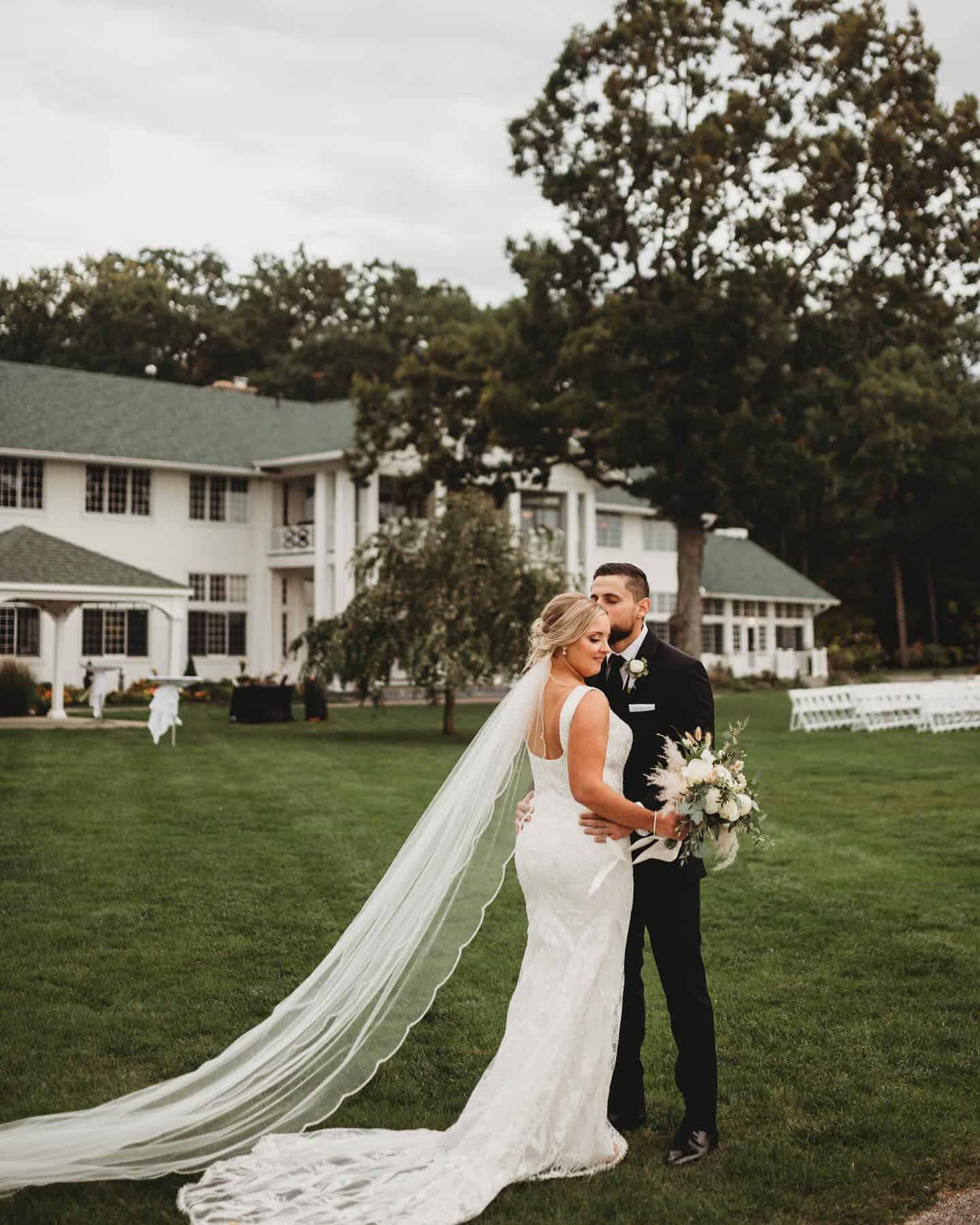 wedding Bride and groom pose together on a lawn in front of a large white house; bride wears a long veil and holds a bouquet, groom wears a black suit.