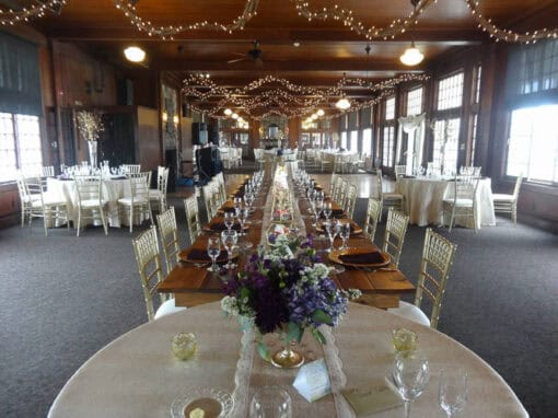 A banquet hall with long and round tables set for an event, decorated with string lights, flowers, glassware, and gold chairs.