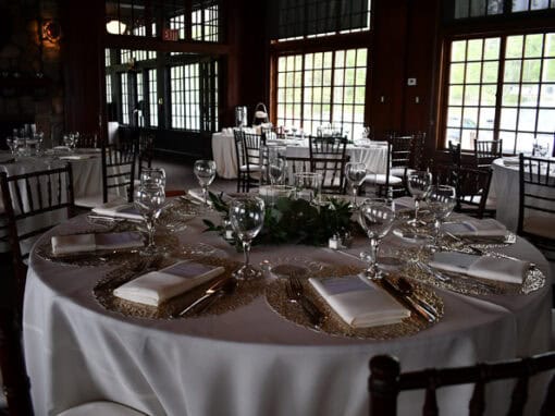 A round dining table is set with white linens, glassware, plates, and napkins in a dimly lit room with large windows and several other arranged tables.