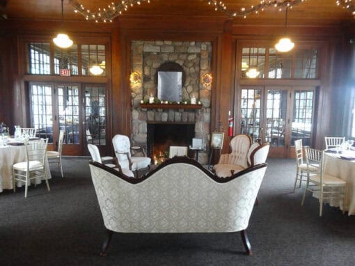 Elegant lounge area with vintage sofas and chairs arranged around a stone fireplace in a wood-paneled room with round dining tables and string lights overhead.