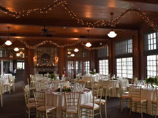 A banquet hall with round tables set for an event, gold chairs, floral centerpieces, and string lights hanging from the wooden ceiling.