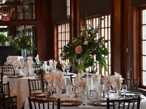 A formal dining room set for an event, featuring round tables with white tablecloths, neatly folded napkins, glassware, and large floral centerpieces.