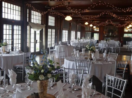 Elegant dining hall decorated for an event with round tables set in white linens, floral centerpieces, and string lights draped across the ceiling.