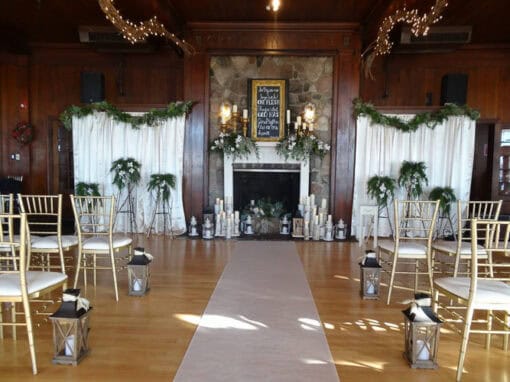 Indoor wedding ceremony at Cromaine Hall, Waldenwoods, with rows of gold chairs, lanterns on the floor, a central aisle, and a fireplace with greenery and candles at the front.