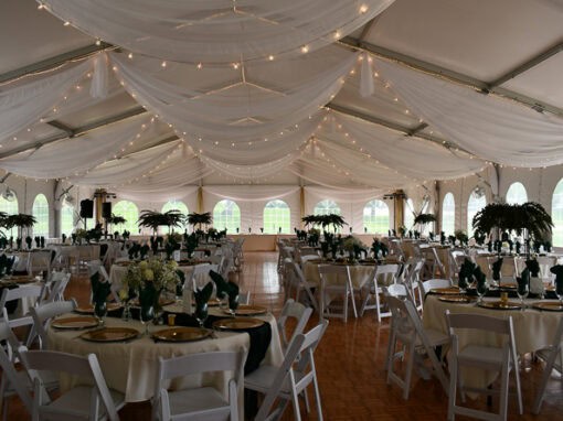 Large wedding tent with draped white ceiling, string lights, round tables set with gold plates, green napkins, white chairs, and a central dance floor.
