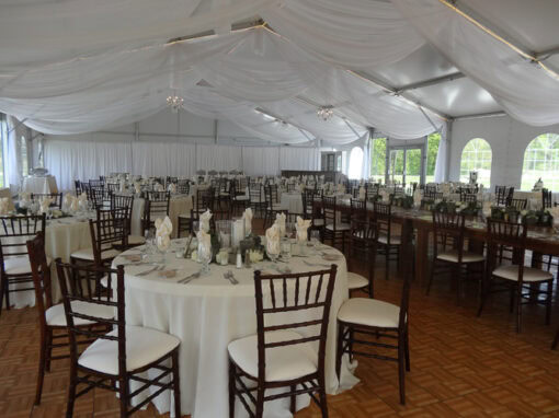A banquet hall with round tables covered in white tablecloths, set with utensils and napkins, and surrounded by wooden chairs under a white draped ceiling.