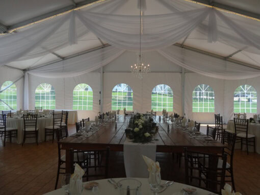A decorated event tent with round and rectangular tables set for a formal meal, white draped ceiling, chandelier, and windows showing a green outdoor area.