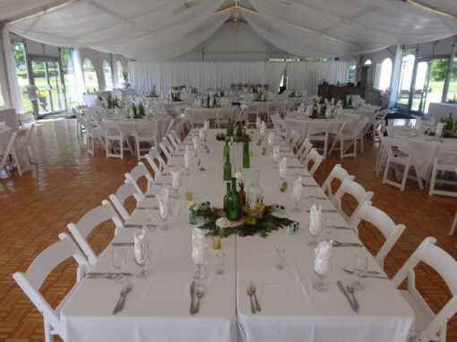 A large white tent is set up for an event with long rectangular tables, white chairs, white tablecloths, and green glass centerpieces.