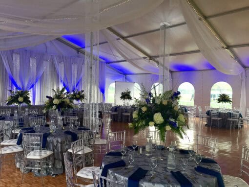 Elegant event venue with round tables, clear chairs, floral centerpieces, and white draping accented by blue lighting inside a large tent with arched windows.