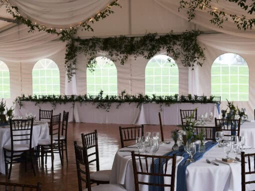 A wedding reception tent with round tables set for guests, white tablecloths, wooden chairs, greenery decorations, and arched windows letting in natural light.