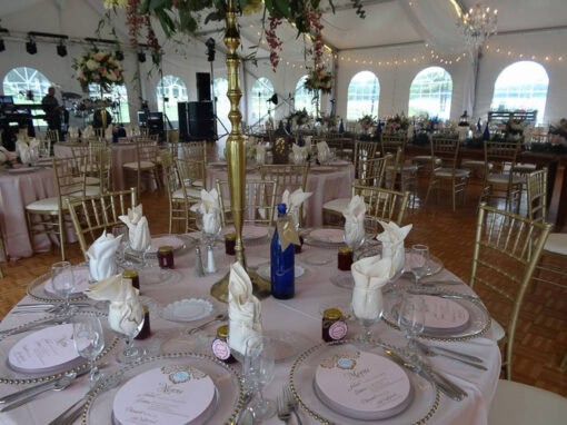Elegant wedding reception setup with round tables, gold chairs, folded napkins, menus, and floral centerpieces inside a decorated tent.