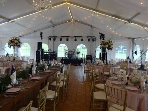 A large wedding reception tent with decorated tables, gold chairs, floral centerpieces, and a stage set up with instruments and lighting equipment.