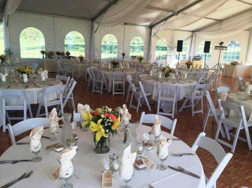 Large white event tent with round tables set for a formal gathering, featuring floral centerpieces, white linens, and folded napkins on each table.