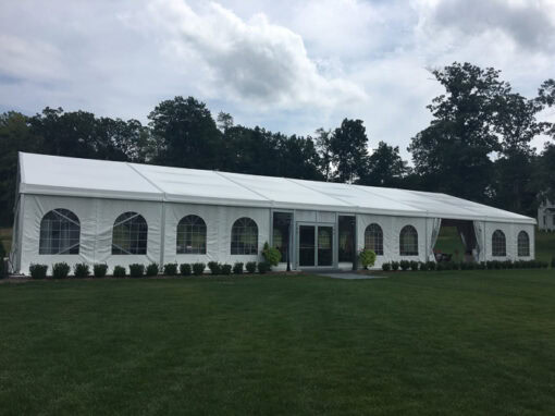 A large white event tent with arched windows stands on a grassy lawn, surrounded by small shrubs and trees in the background.