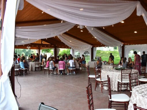 Group seated at round tables under an outdoor pavilion with white drapes and string lights during an event at Sunshine Cove at Waldenwoods.