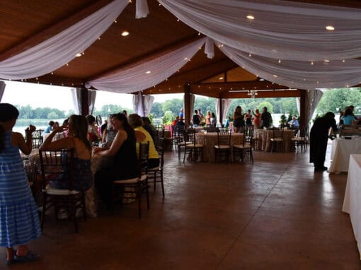 Guests sit at tables under a pavilion with white drapes and string lights for an outdoor event, with trees in the background at Waldenwoods' Sunshine Cove.