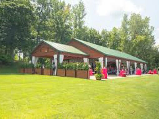 Sunshine Cove; a wooden pavilion with a green roof on a grassy lawn, surrounded by trees, with red tablecloth-covered tables set up outside.