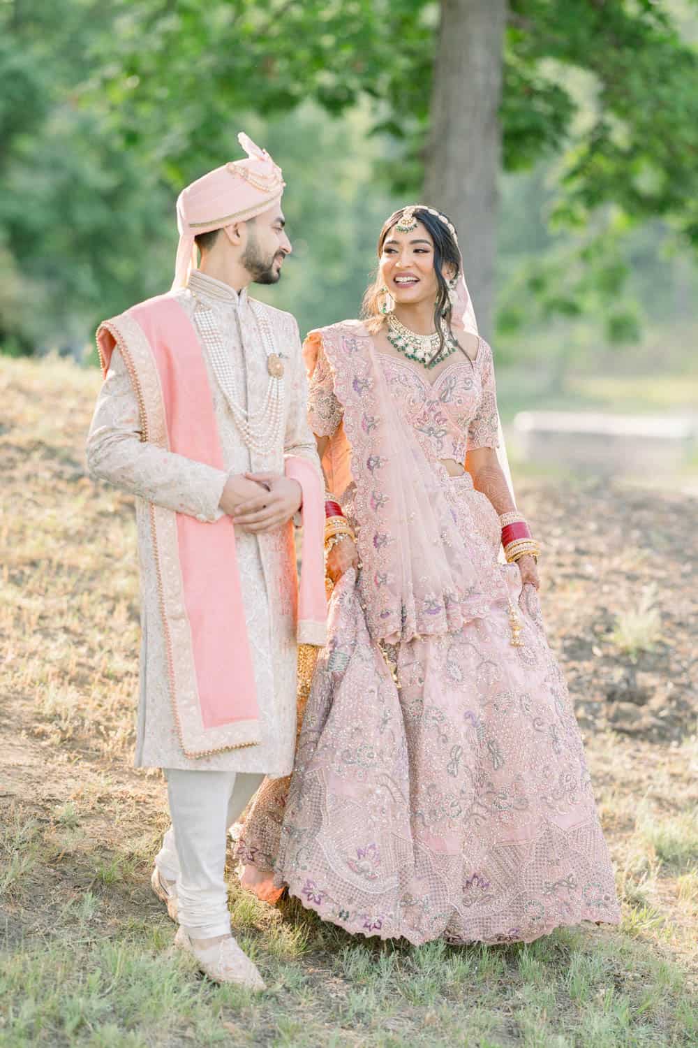 Bride and groom in traditional South Asian attire walk outdoors smiling; she wears a pink lehenga, he wears a cream sherwani with pink stole and turban.