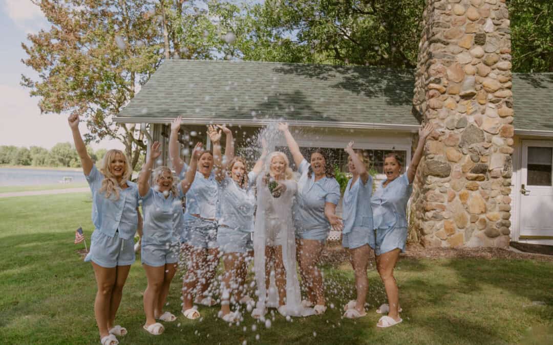 Group of women in matching light blue pajamas celebrate outdoors at Waldenwoods as the bride sprays champagne; they stand on grass near a chimney and house.