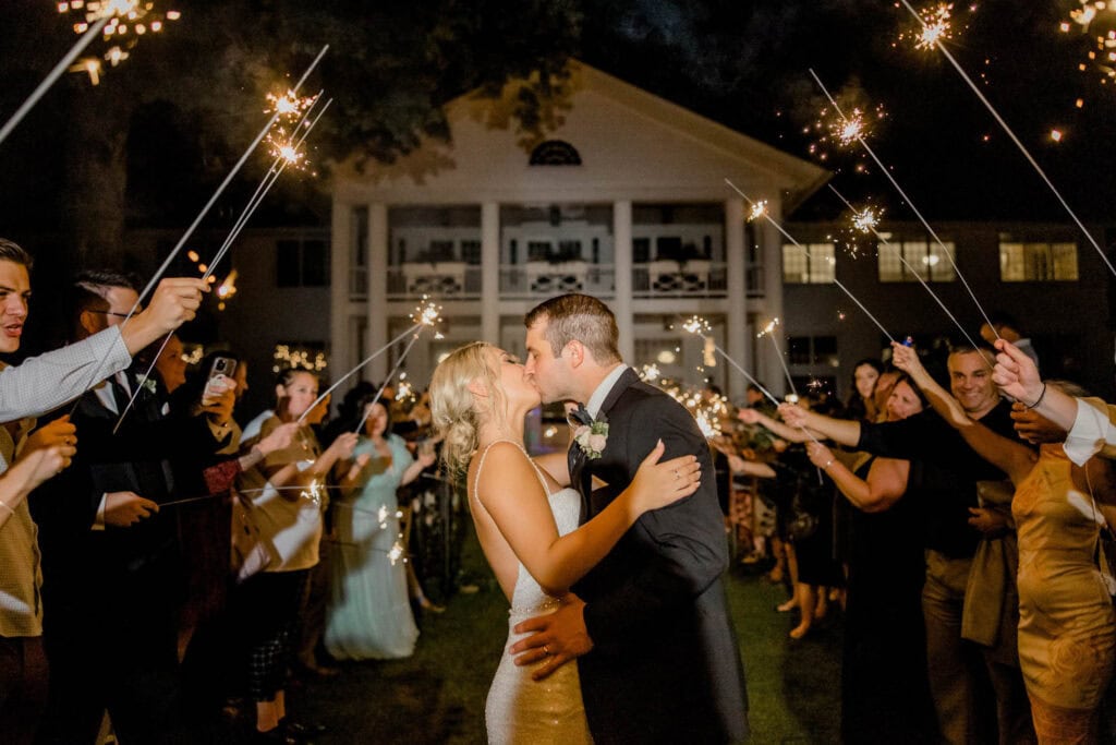 A bride and groom kiss outdoors at night, surrounded by guests holding sparklers in front of a large white building.