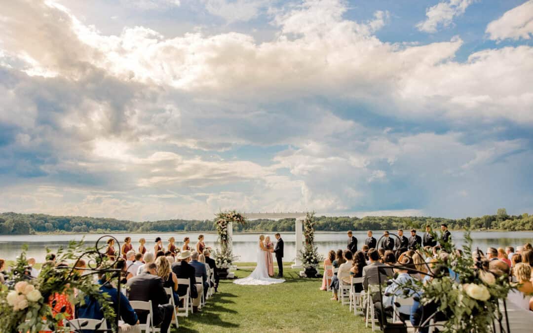 A bride and groom stand at an outdoor altar by a lake, surrounded by guests seated in rows, with floral decorations and a partly cloudy sky overhead.