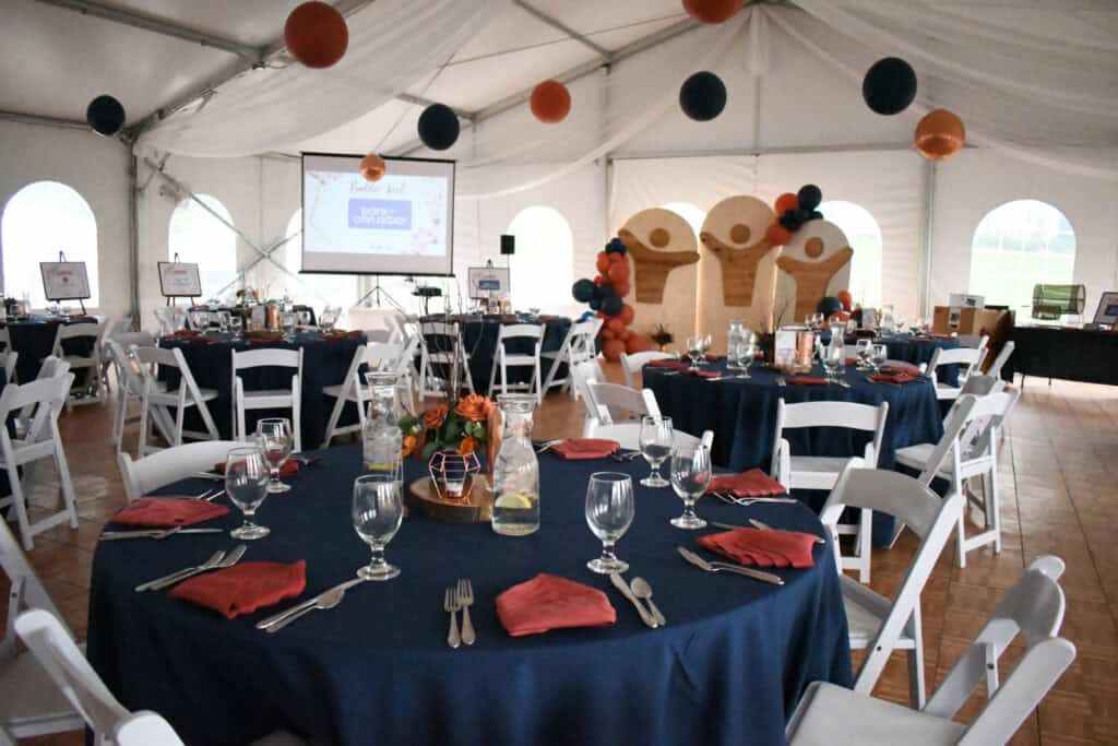 Decorated event tent at Waldenwoods with round tables in navy tablecloths, red napkins, white chairs, orange and navy balloon decorations, and a projector screen in the background.