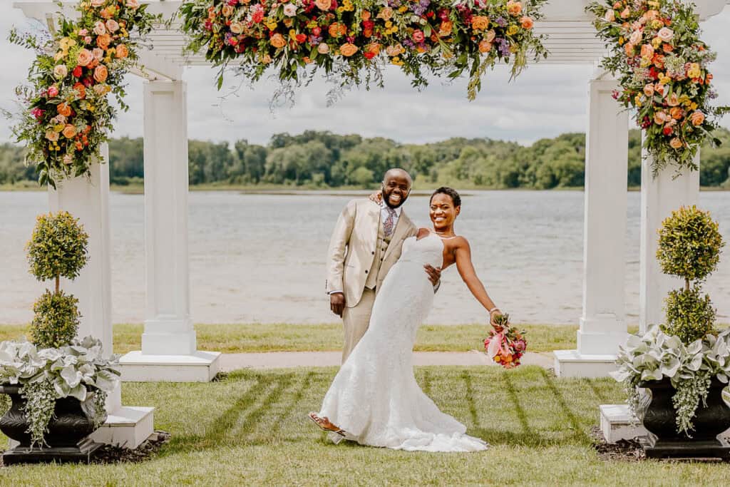 A bride and groom pose and smile under a floral arch outdoors near a lake, with greenery and trees in the background.