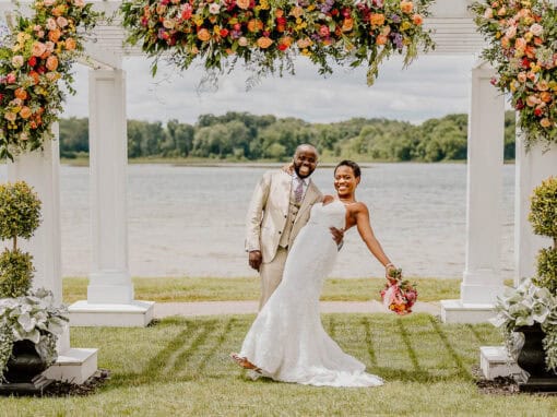 A bride and groom pose and smile under a floral arch outdoors near a lake, with greenery and trees in the background.