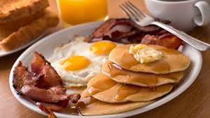 A breakfast plate with pancakes topped with butter and syrup, fried eggs, bacon, toast, a glass of orange juice, and a cup of coffee.